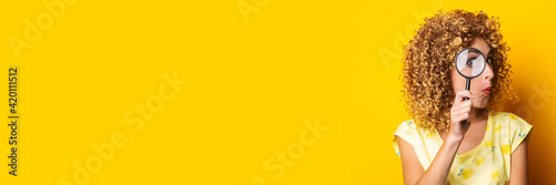 surprised curly young woman looks through a magnifying glass on a yellow background. Banner.