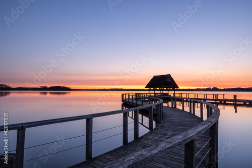 Curvy Pier construction and shelter with thatched roof at beautiful colorful sunrise under clear sky at Lake Hemmelsdorf, Schleswig-Holstein, Northern Germany