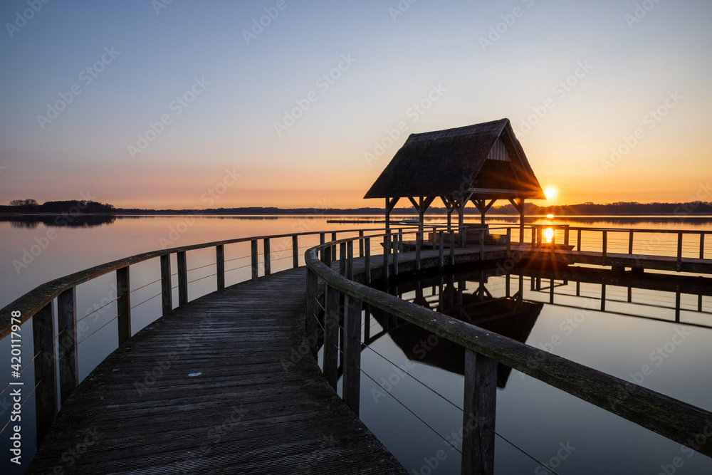 Fototapeta premium Swinging Pier construction and shelter with thatched roof with bright sunsatr at beautiful colorful sunrise under clear sky at Lake Hemmelsdorf, Schleswig-Holstein, Northern Germany
