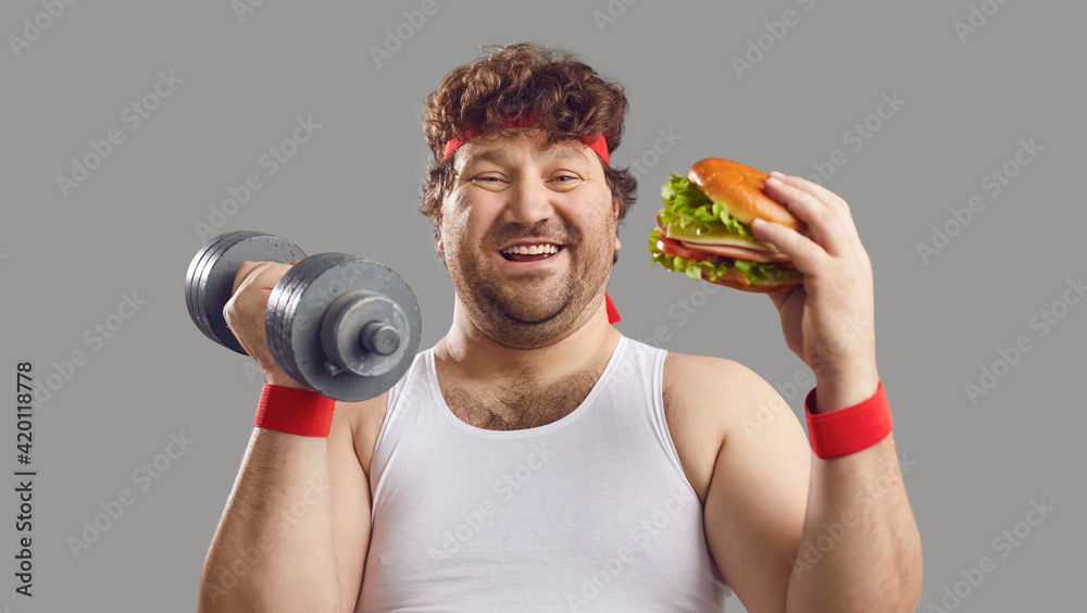 Funny smiling man with dumbbell and delicious burger looking at camera.  Happy fat guy holding free weights and eating big yummy hamburger. Sport,  food, failed diet, workout exercise, cheat day concept Stock