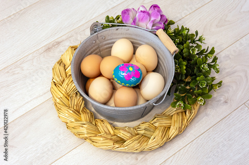 Metal bucket with chicken eggs and a colorful, hand-painted Easter egg. Easter composition with flowers and straw wreath on a wooden background. 