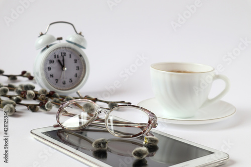 Glasses for vision lie on the tablet against the background of a cup of tea and an alarm clock, willow branches, a white background-the concept of spending time on the Internet by older people