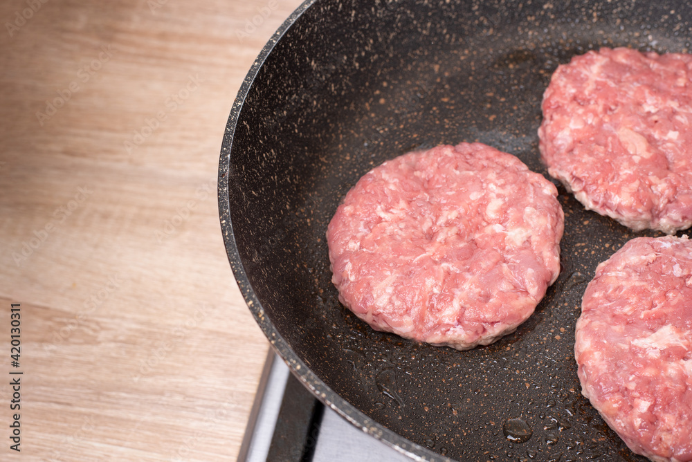 Three raw beef patties grilling in a non-stick frying pan in the kitchen. Three raw beef patties grilling in a non-stick frying pan in the kitchen.