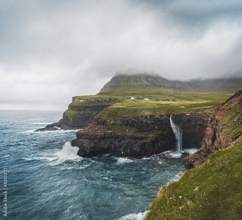 Gasadalur village and Mulafossur its iconic waterfall, Vagar, Faroe Islands, Denmark. Rough see in the north atlantic ocean. Lush greens during summer.