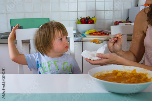 Mom feeding her disabled child with down syndrome. Mother giving food to toddler at home. Child doesn't like food.
