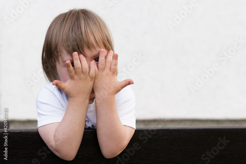 Horizontal outdoor image of little boy with down syndrome covering his eyes with both hands, playing hide and seek  leaned on fance. Adorable child having fun outdoors on terrace.