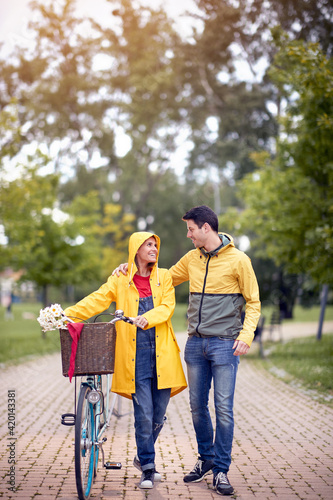 vertical image of beautiful female looking with love and affection young male hugging her while pushing the bicycle on a rainy day in park