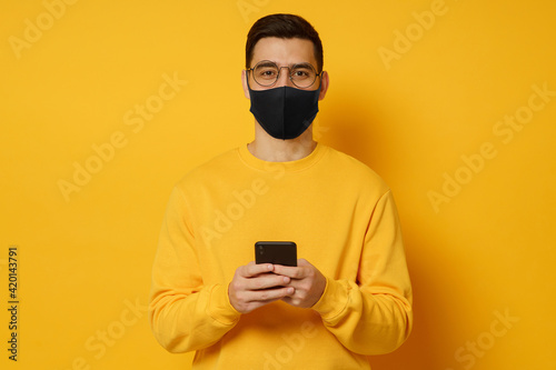 Guy wearing black covid mask, standing in front of camera with smartphone in hands,  isolated on yellow background © Damir Khabirov