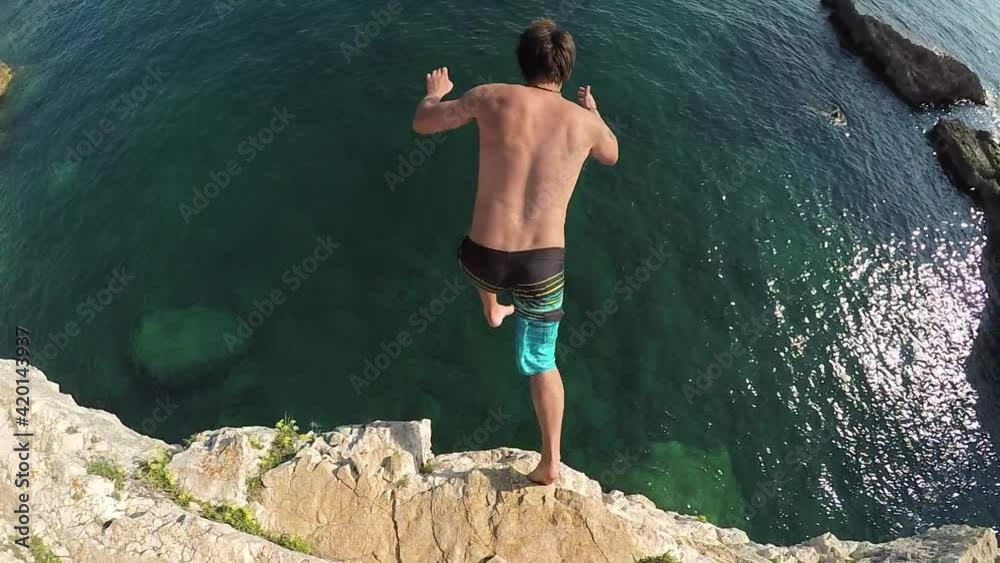 SLOW MOTION, UNDERWATER: Active young man jumping off a high dangerous rocky cliff into beautiful crystal clear ocean on amazing sunny summer day. Sea foaming, water drops splashing high in the air