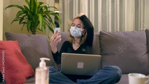 Young Turkish woman with a facial mask, having a video call on her smartphone while sitting on the sofa. There is a laptop computer on her lap, coffee cup and a hand sanitizer bottole on the table. photo