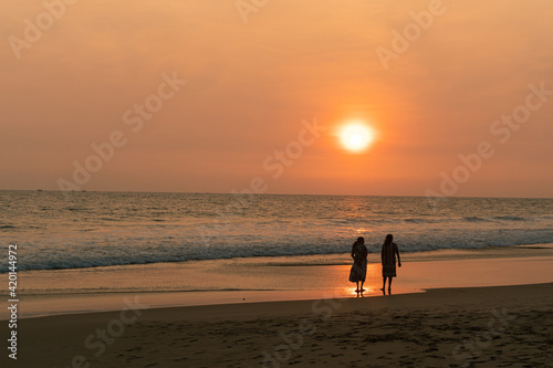 Kobiety spacerujące brzegiem oceanu o zachodzie słońca, ciemno pomarańczowy nastrojowy krajobraz.