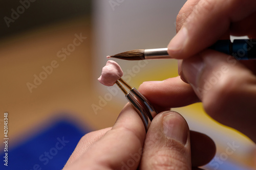 Dental technician prepares to apply ceramic material to a dental implant crown in a dental laboratory using tweezers and a brush. Denture manufacturing process close up. Modern dental technology.