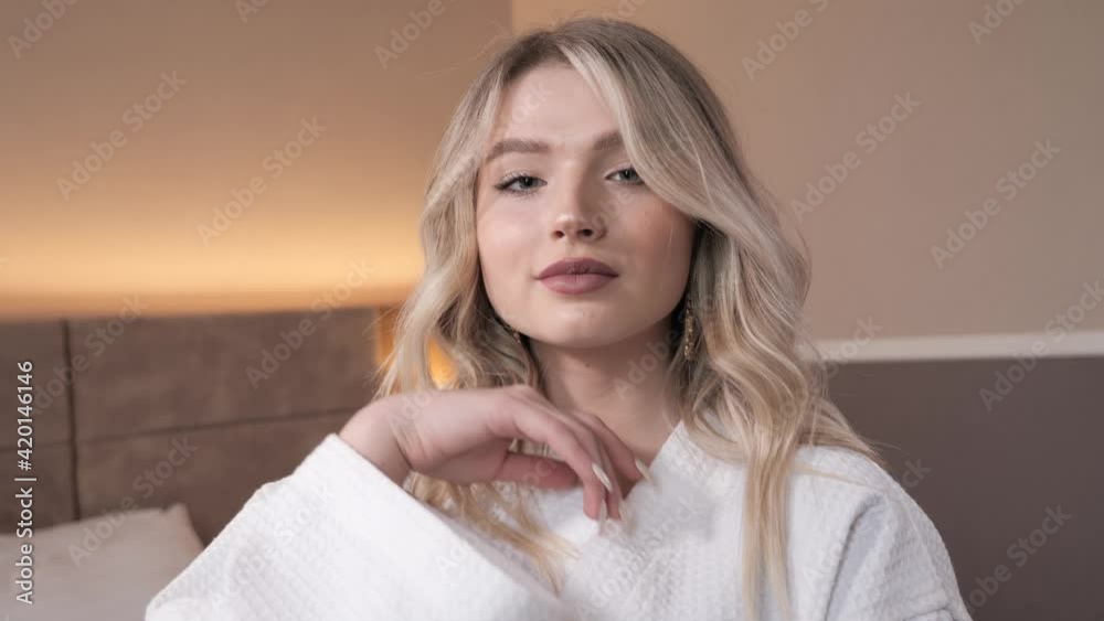 Beauty shooting of young caucasian female model with blonde wavy hair natural makeup wearing white cotton bathrobe smiling looking to camera touching her face with her fingers feeling comfort cosiness Beauty shooting of young caucasian female model with blonde wavy hair natural makeup wearing white cotton bathrobe smiling looking to camera touching her face with her fingers feeling comfort cosiness