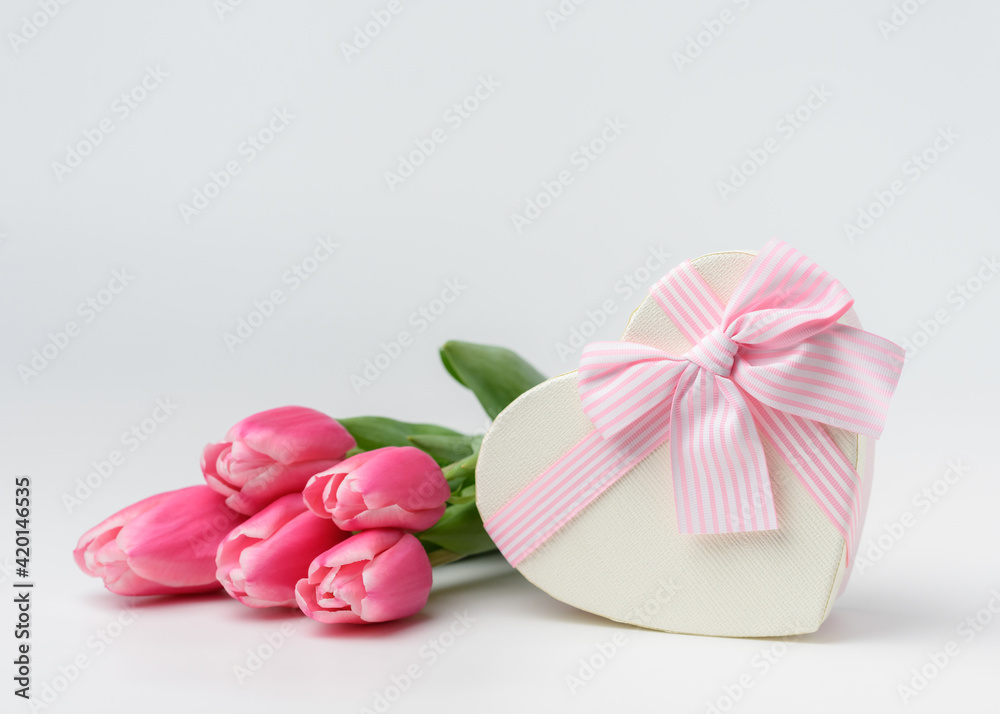 Beautiful tulips of pink color in a silk ribbon near a heart-shaped gift box with a ribbon and a beautifully tied pink bow. The concept of a gift and surprise for a loved one.