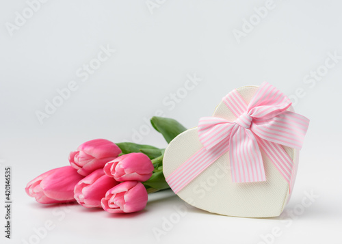 Beautiful tulips of pink color in a silk ribbon near a heart-shaped gift box with a ribbon and a beautifully tied pink bow. The concept of a gift and surprise for a loved one.