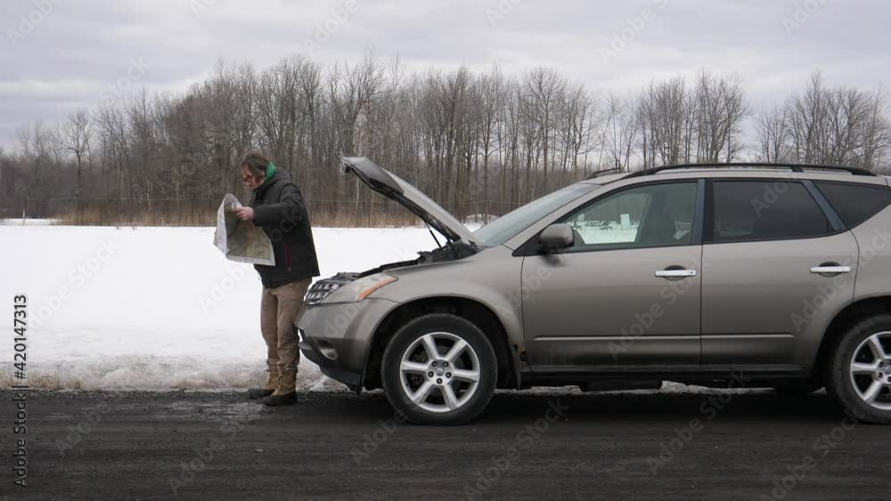 Man with broke down car on the side of the road during winter looking at the map to find the nearest mechanic shop Man with broke down car on the side of the road during winter looking at the map to find the nearest mechanic shop