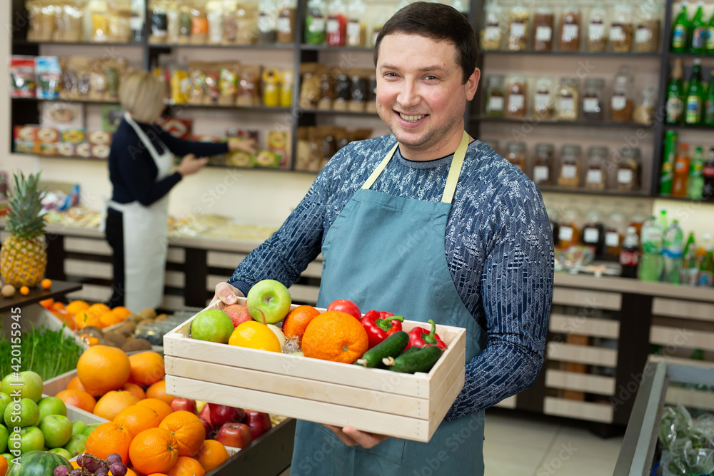 Smiling male salesman holds a wooden box with vegetables and fruits in the store.