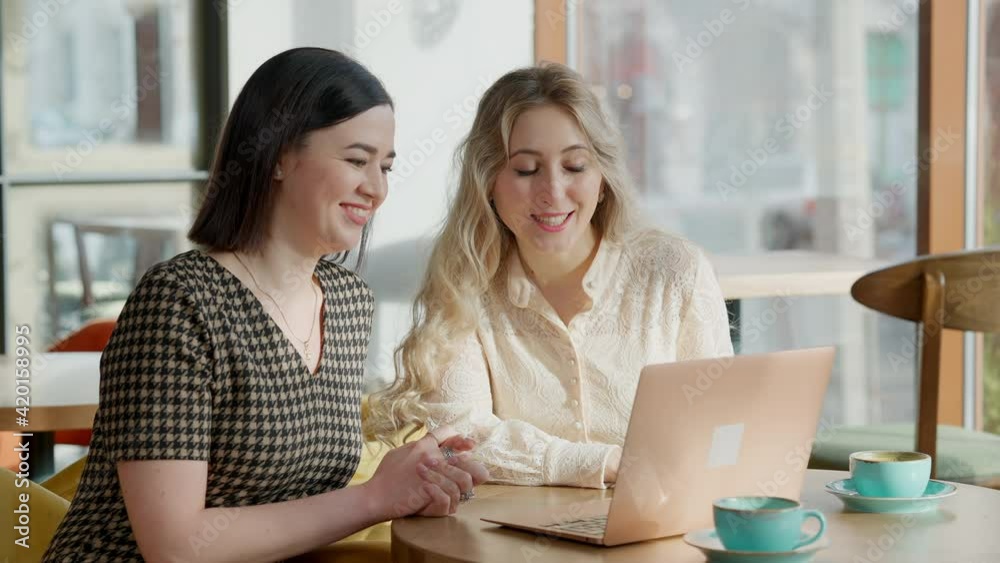 Two positive women using social media on laptop and talking sitting in cafe. Portrait of smiling elegant friends surfing internet on lunch break in restaurant. Modern lifestyle concept. Two positive women using social media on laptop and talking sitting in cafe. Portrait of smiling elegant friends surfing internet on lunch break in restaurant. Modern lifestyle concept.