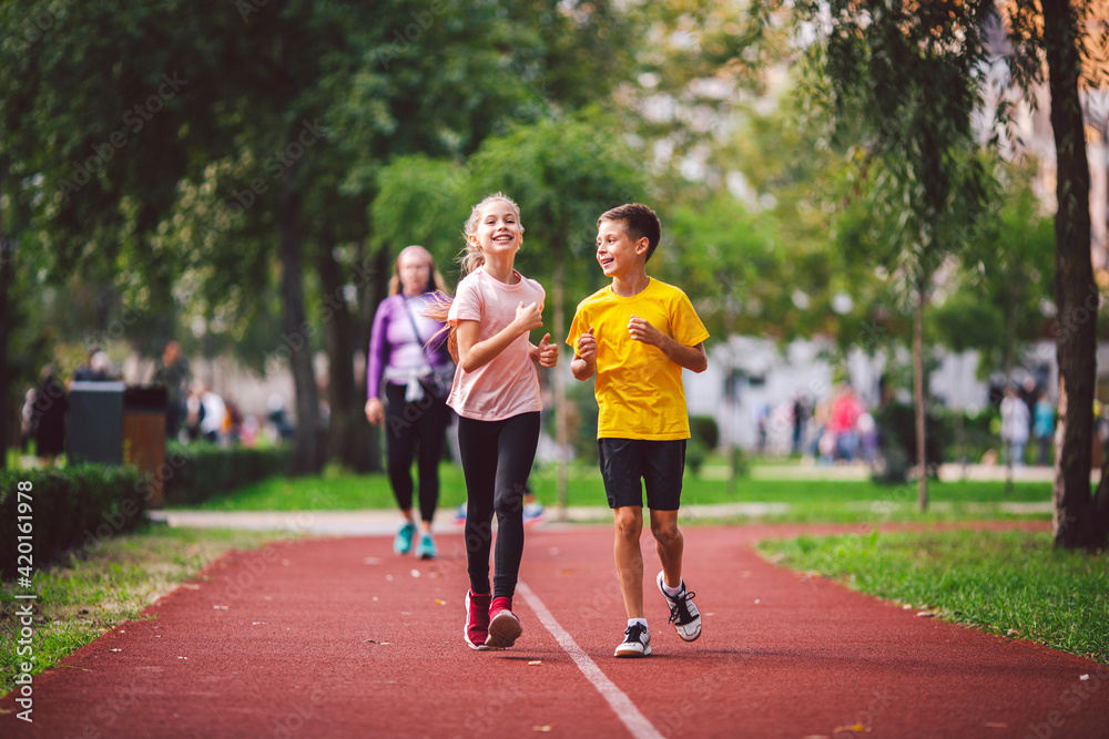 Active recreation and sports children in pre-adolescence. Caucasian twins boy and girl 10 years old jogging on red rubber track through park. Children brother and sister running on treadmill outside