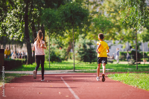 Couple of kids boy and girl doing cardio workout, jogging in park on jogging track red. Cute twins runing together. Run children, young athletes. Teen brother and sister running along path outdoors