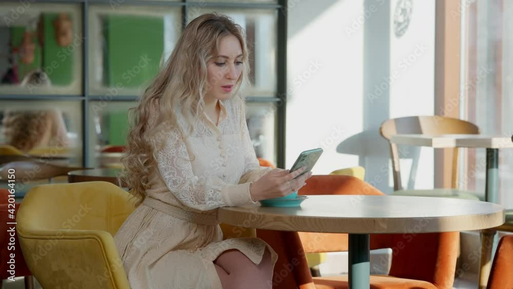 Slim happy relaxed blond woman surfing social media on smartphone drinking coffee in restaurant. Portrait of positive Caucasian millennial enjoying morning in cafe indoors. Slim happy relaxed blond woman surfing social media on smartphone drinking coffee in restaurant. Portrait of positive Caucasian millennial enjoying morning in cafe indoors.