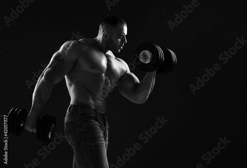 Muscular men is working out in gym, lifting alternately two big heavy dumbbells, doing exercises for biceps over black background with copy space. Young man lifting weights. Black and white  © Dmitry Lobanov
