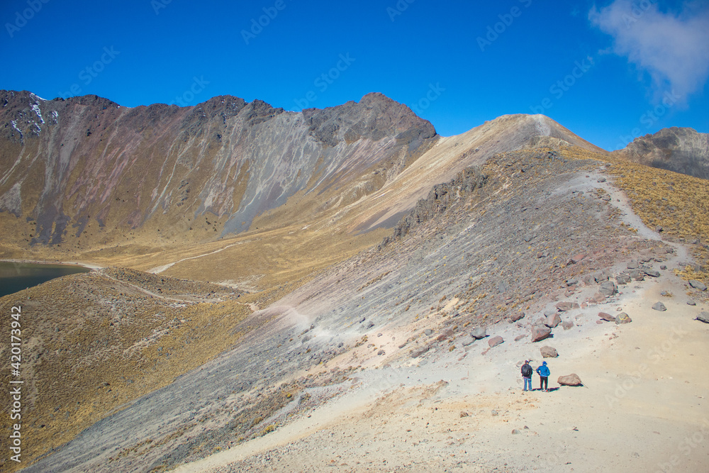 Fototapeta premium Two adult men walking on the rocky path of a snowy mountain in the middle of a cold summer morning with blue sky.