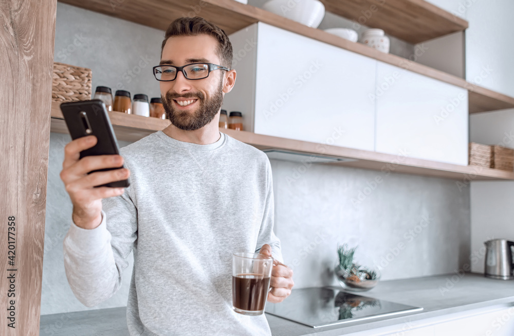 smiling man with smartphone and Cup of coffee standing in his kitchen smiling man with smartphone and Cup of coffee standing in his kitchen