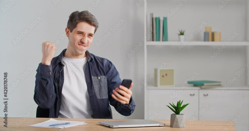 Caucasian male businessperson millennial freelancer sitting at desk finishing up phone call selling online using smartphone making victory gesture receiving good news rejoicing over success triumph Caucasian male businessperson millennial freelancer sitting at desk finishing up phone call selling online using smartphone making victory gesture receiving good news rejoicing over success triumph