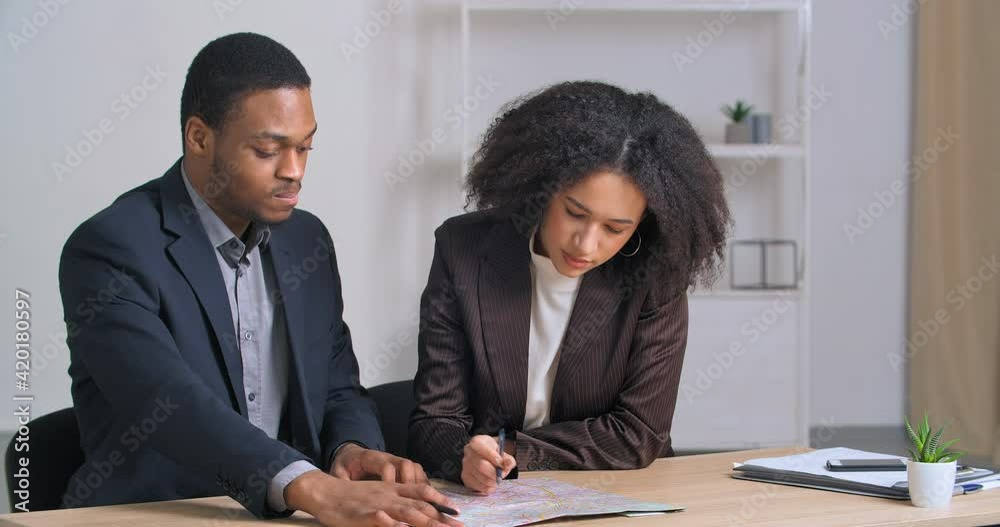 Couple of colleges afro american businessman and woman architect two designers analyzing plan looking at diagram studying map pointing with hand in document sitting at office table discussing project Couple of colleges afro american businessman and woman architect two designers analyzing plan looking at diagram studying map pointing with hand in document sitting at office table discussing project