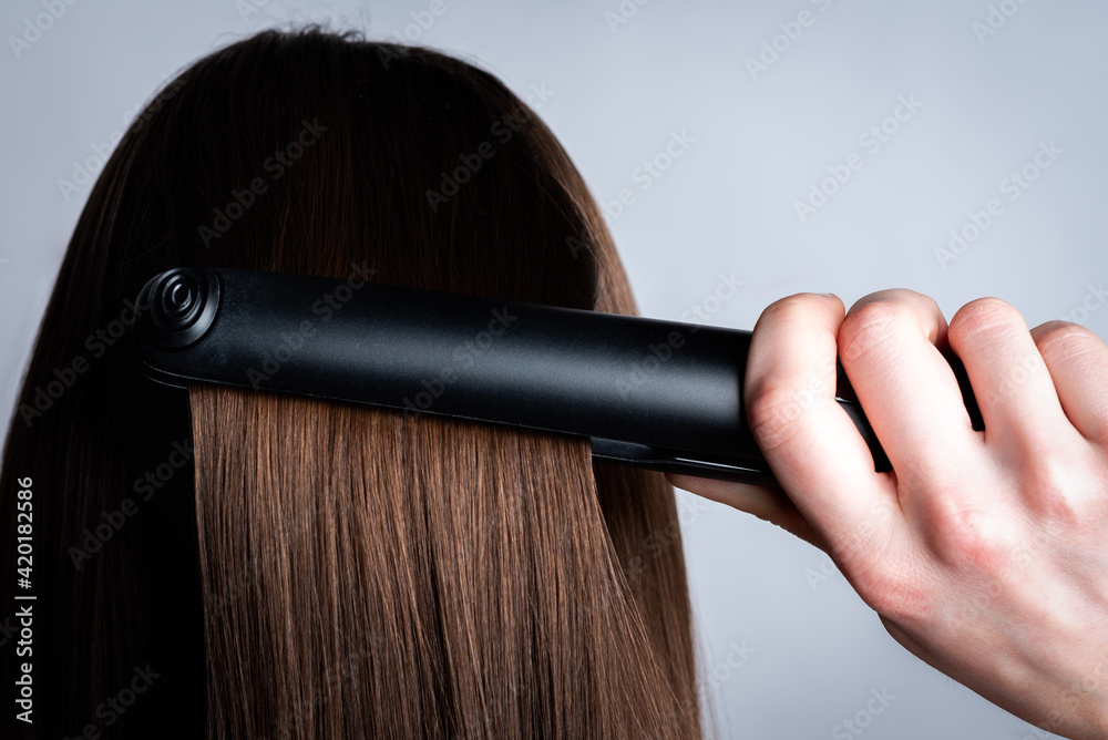 Fototapeta premium Young girl is doing her hair using a straightener on gray background.