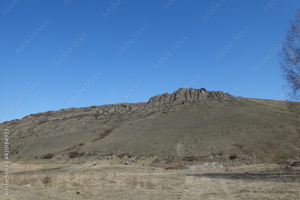 A field and a rocky mountain in early spring against a blue sky. A gray and bleak spring landscape in the countryside.
