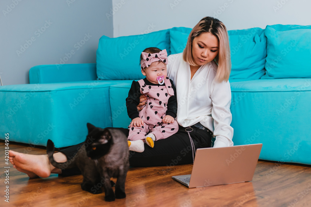 A young mother works over the Internet. A woman of Asian appearance uses a laptop, holding a baby in her arms, sitting on the floor next to a cat.