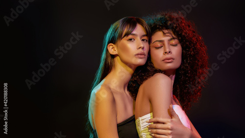 Two stylish young women with professional art makeup posing together in neon light isolated over black background