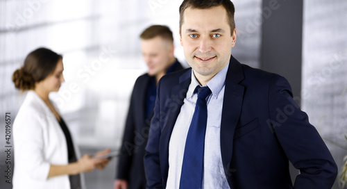 Portrait of a self-confident middle aged businessman in a blue suit  standing in a modern office with his colleagues at the background. Concept of business success