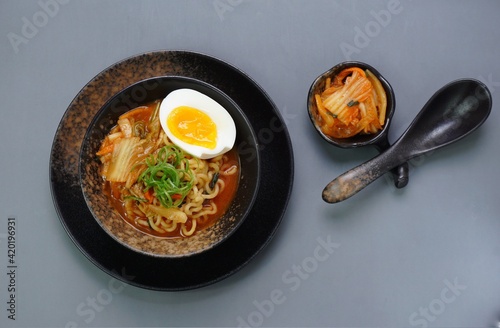 Top view of instan ramyeon or Korean noodles soup on black bowl, topped with half boiled egg, leek and Kimchi on small bowl. Isolated gray background with spoon, napkin and wooden board