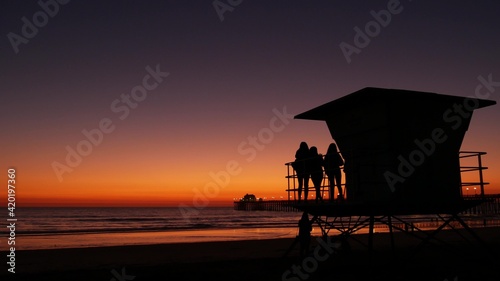 Young teen girls silhouettes near lifeguard tower, friends on pacific ocean beach, sunset dusk in Oceanside, California USA. Unrecognizable teenagers, people and twilight gradient purple violet sky. © Dogora Sun