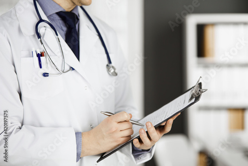 Unknown male doctor standing and working with clipboard of medication history records in clinic at his working place  closeup. Perfect medical service in hospital. Medicine and healthcare concept