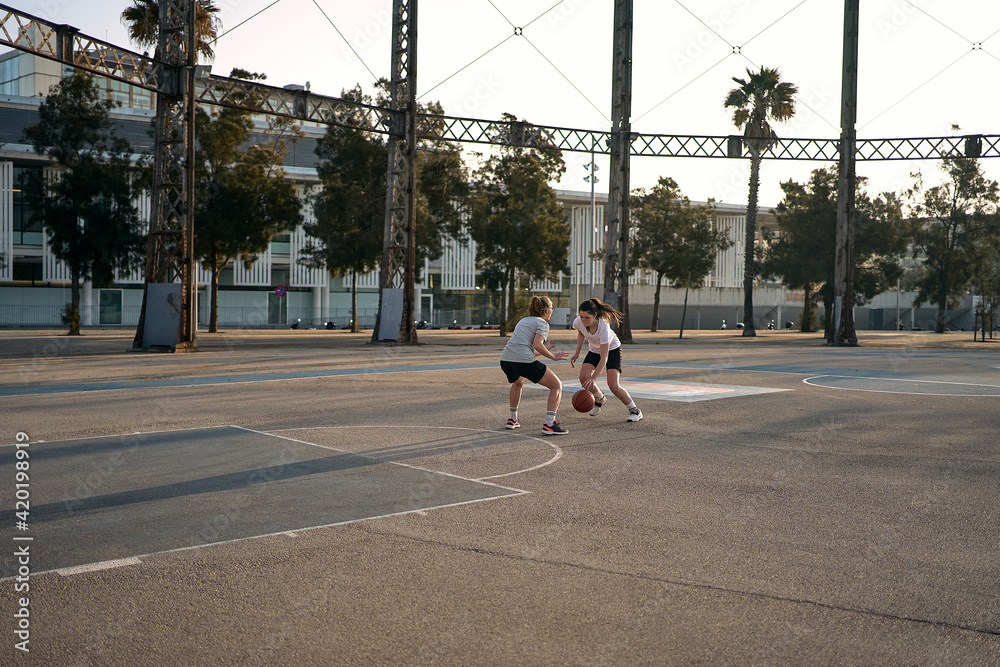 Player dribbling her opponent to make a point on an urban court surrounded by palm trees on a sunny morning.