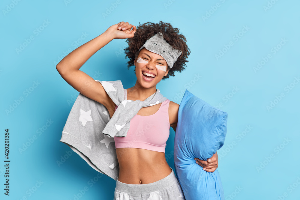 Indoor shot of joyful Afro American woman has fun before going to bed dances carefree holds soft pillow dressed in casual pajama tilts head applies beuty pads under eyes isolated over blue wall