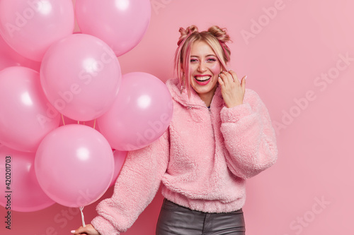 Happy beautiful woman has two buns dressed in fur coat smiles gladfully wears makeup holds bunch of inflated balloons enjoys partying and celebration isolated over pink background. Monochrome