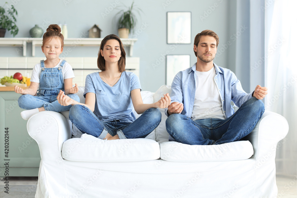 Calm young family with little daughter sit on couch practice yoga together, happy parents with small preschooler girl child rest on sofa meditate relieve negative emotions on weekend at home