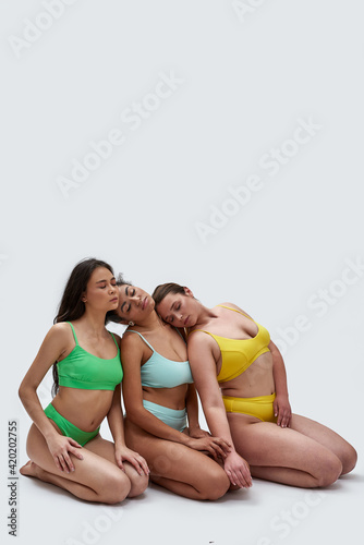 Full length shot of three diverse young women in colorful underwear posing together  leaning on each other  sitting on knees with eyes closed isolated over white background