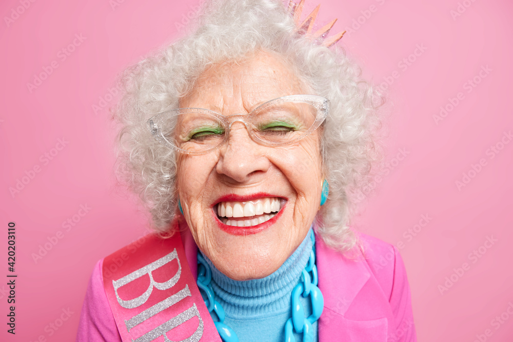 Close up shot of positive wrinkled old woman smiles toothily at camera wears transparent glasses princess crown on head stylish outfit applies bright makeup expresses joy isolated over pink background Close up shot of positive wrinkled old woman smiles toothily at camera wears transparent glasses princess crown on head stylish outfit applies bright makeup expresses joy isolated over pink background