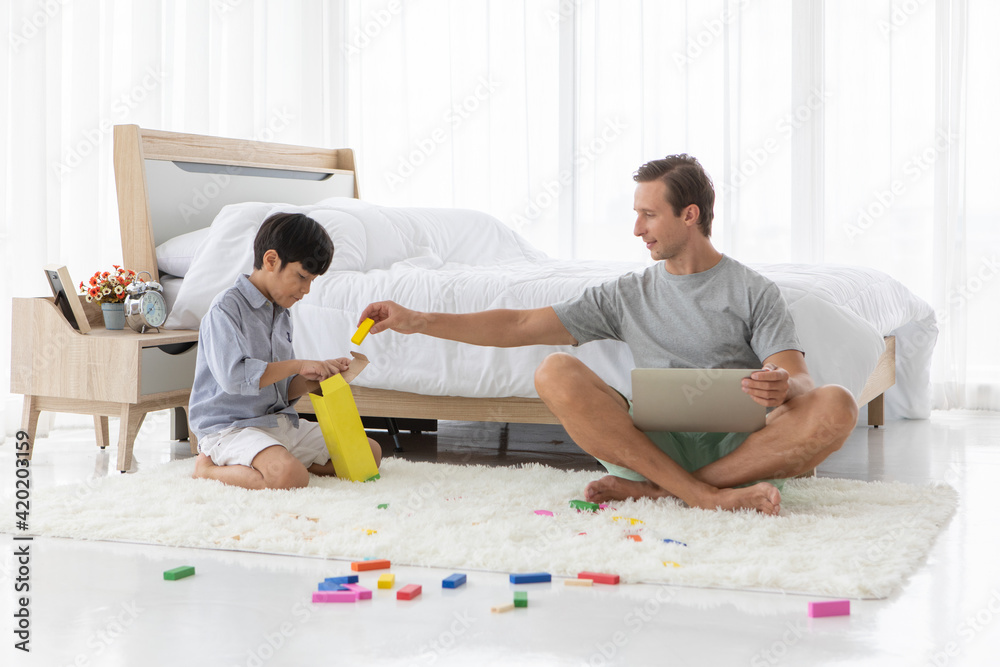 Father sitting andworking with notebook computer while his mixed-race son playing colorful wooden block in home bedroom. Idea for lifestyle of working from home Father sitting andworking with notebook computer while his mixed-race son playing colorful wooden block in home bedroom. Idea for lifestyle of working from home