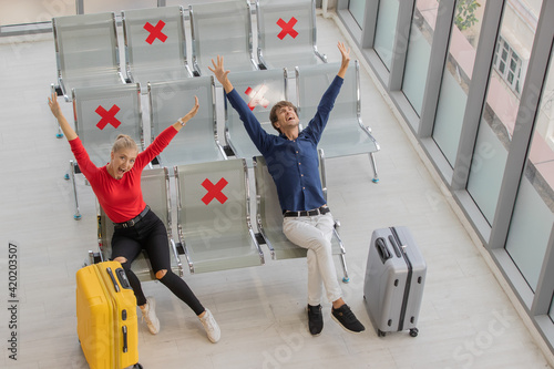 Young man and woman lovers travelers sitting row seats in airport lobby with social distancing sign and rise hands for cheer up. New normal traveling concept. photo