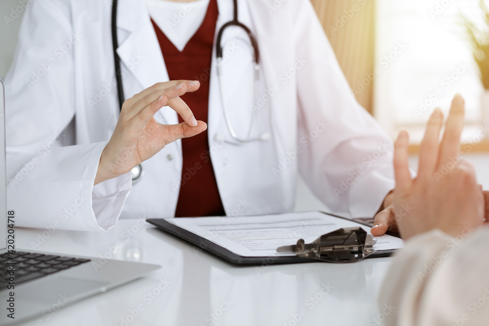 Unknown woman-doctor and patient discussing something while sitting at the table in sunny clinic, close-up Unknown woman-doctor and patient discussing something while sitting at the table in sunny clinic, close-up