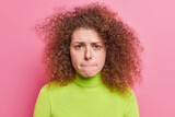 People human reactions and emotions concept. Worried dissatisfied curly haired young European woman wants to cry presses lips dressed in casual green turtleneck isolated over pink background.
