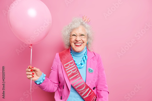 Isolated shot of beautiful wrinkled woman smiles positively celebrates anniversary has fun on party for elderly people wears festive clothes with ribbon holds inflated balloon enjoys birthday © wayhome.studio 