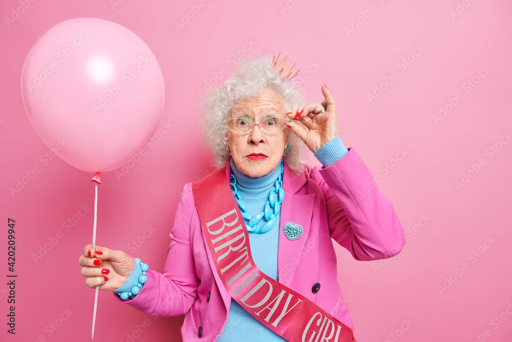 Photo of wondered wrinkled woman keeps hand on rim of spectacles hears surprising news dressed in fashionable outfit holds inflated balloon celebrates birthday isolated over pink background. Photo of wondered wrinkled woman keeps hand on rim of spectacles hears surprising news dressed in fashionable outfit holds inflated balloon celebrates birthday isolated over pink background.